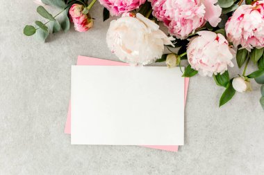 Sahte davetiye, boş kağıt tebrik kartı, pembe zarf ve gri taş masada şakayıklar. Çiçek arkaplanı. Düz yatış, üst görünüm.