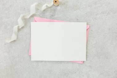 Sahte davetiye, boş kağıt tebrik kartı, pembe zarf ve gri taş masada şakayıklar. Çiçek arkaplanı. Düz yatış, üst görünüm.