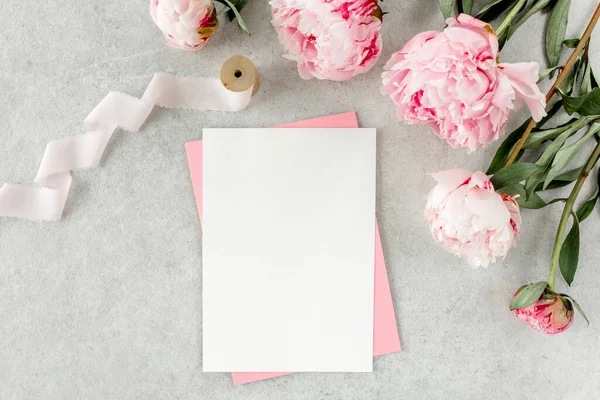 Sahte davetiye, boş kağıt tebrik kartı, pembe zarf ve gri taş masada şakayıklar. Çiçek arkaplanı. Düz yatış, üst görünüm.