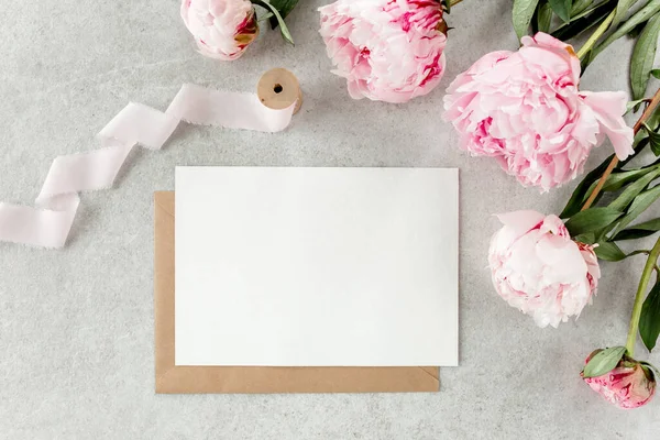 Sahte davetiye, boş kağıt tebrik kartı ve gri taş masadaki şakayıklar. Çiçek arkaplanı. Düz yatış, üst görünüm.
