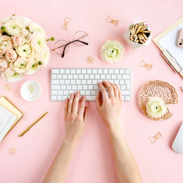 Stilisierter, pinkfarbener Home-Office-Schreibtisch für Frauen. Arbeitsplatz mit weiblichen Händen, Computer, Strauß Hahnenfuß und Rosen, Klemmbrett auf rosa Hintergrund. Flache Lage. — Stockfoto