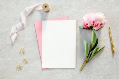 Sahte davetiye, boş kağıt tebrik kartı, pembe zarf ve gri taş masada şakayıklar. Çiçek arkaplanı. Düz yatış, üst görünüm.