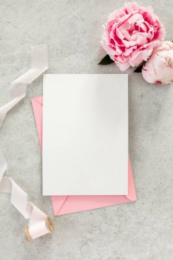 Sahte davetiye, boş kağıt tebrik kartı, pembe zarf ve gri taş masada şakayıklar. Çiçek arkaplanı. Düz yatış, üst görünüm.