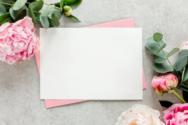 Sahte davetiye, boş kağıt tebrik kartı, pembe zarf ve gri taş masada şakayıklar. Çiçek arkaplanı. Düz yatış, üst görünüm.