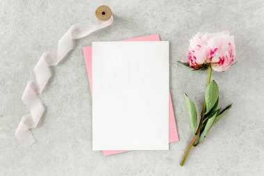 Sahte davetiye, boş kağıt tebrik kartı, pembe zarf ve gri taş masada şakayıklar. Çiçek arkaplanı. Düz yatış, üst görünüm.