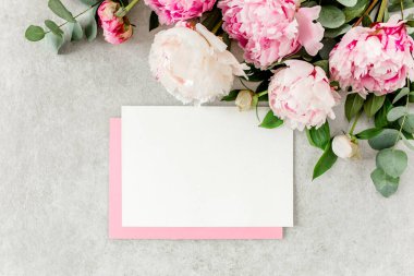 Sahte davetiye, boş kağıt tebrik kartı, pembe zarf ve gri taş masada şakayıklar. Çiçek arkaplanı. Düz yatış, üst görünüm.