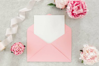 Sahte davetiye, boş kağıt tebrik kartı, pembe zarf ve gri taş masada şakayıklar. Çiçek arkaplanı. Düz yatış, üst görünüm.