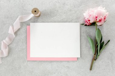 Sahte davetiye, boş kağıt tebrik kartı, pembe zarf ve gri taş masada şakayıklar. Çiçek arkaplanı. Düz yatış, üst görünüm.