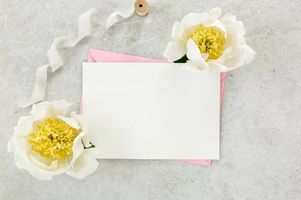 Mockup invitation, blank paper greeting card, pink envelope and peonies on gray stone table. Flower background. Flat lay, top view. — Stock Photo, Image