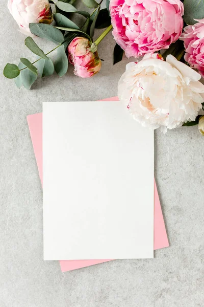 Invitation maquette, carte de vœux en papier vierge, enveloppe rose et pivoines sur table en pierre grise. Fond de fleur. Couché plat, vue du dessus. — Photo