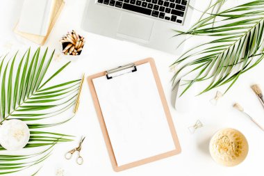 Ev ofisinin masası düzdü. Bilgisayarı, panosu, tropik palmiye yaprakları, beyaz arka planda aksesuarları olan bir kadın çalışma alanı. Üst görünüm. 