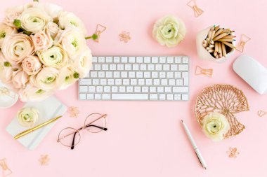 Tarz sahibi, pembe kadınların ofis masası. Bilgisayarlı çalışma alanı, buket ranunculus ve güller, pembe arka planda pano. Düz yatıyordu. Üst görünüm.