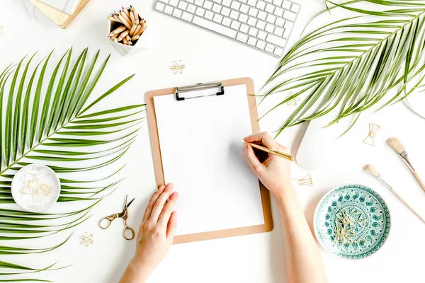 Female workspace with female hands, clipboard, tropical palm leaves, computer, accessories on white background. Stylized womens desk. Flat lay. Top