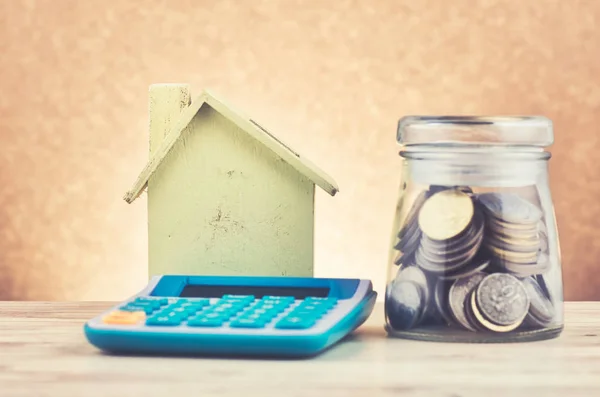 Tarro de vidrio con monedas, calculadora y réplica de la casa en el escritorio de madera para el concepto de préstamos hipotecarios — Foto de Stock