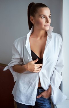 Pencereden bakarken düğmeleri iliklenmemiş bir kadın gülümsüyor..