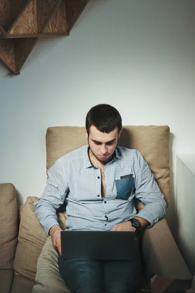 Atractivo hombre con una camisa y vaqueros sentados en el intside de la habitación y trabajando en el ordenador portátil — Foto de Stock