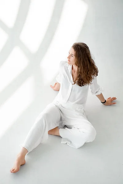 Νεαρή γυναίκα με λευκό πουκάμισο σε λευκό φόντο. Όμορφη κοπέλα με άσπρα ρούχα κάθεται στο πάτωμα σε λευκό φόντο. — Φωτογραφία Αρχείου