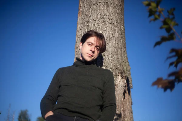 The guy is standing by a tree. A young man in a half-dress.