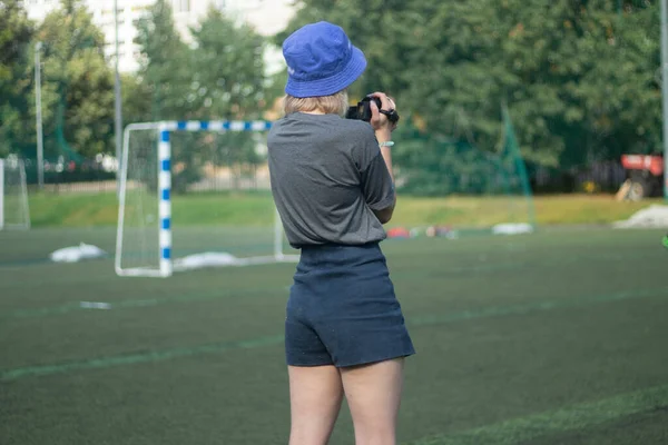 La chica toma fotos de deportes. La chica está filmando un partido de fútbol. Chica en pantalones cortos en el patio de recreo. —  Fotos de Stock