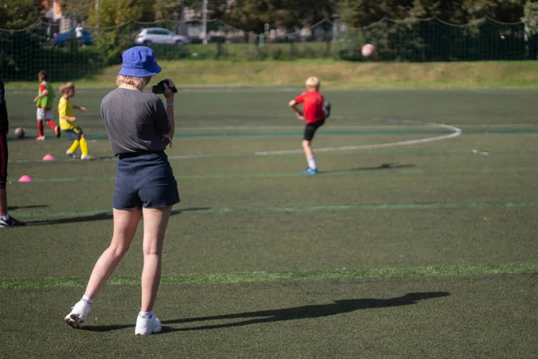 A menina tira fotos de esportes. A rapariga está a filmar um jogo de futebol. Menina em shorts no parque infantil. — Fotografia de Stock