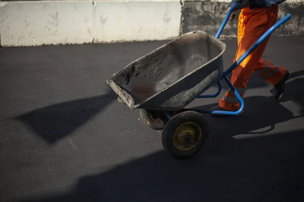 Robotnik niesie wózek. Naprawa dróg. Ładowarka ciągnie złamany asfalt w wózku jednokołowym.. — Zdjęcie stockowe