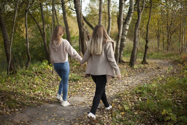 Flickorna håller varandra i handen. Vänner går i höstskogen. Två flickor går glatt genom parken. — Stockfoto