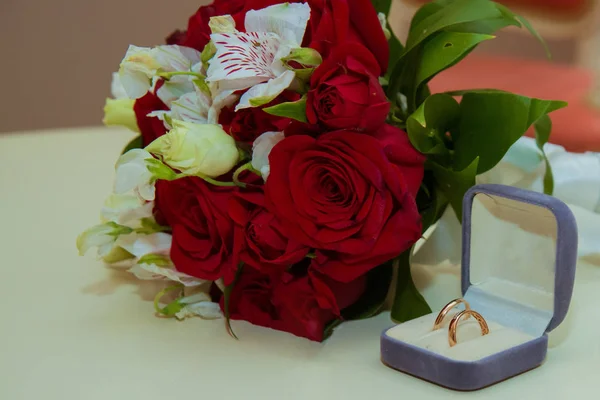 Wedding ring. Two gold vintage rings of the bride and groom with diamonds are in a blue box and a bouquet of red roses and white flowers — Stock Photo, Image