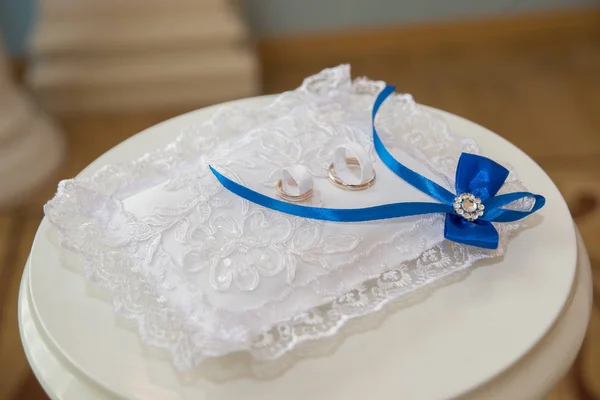 Piękne obrączki leżą na poduszce z białą koronką i niebieskimi wstążkami w kształcie łuku. — Zdjęcie stockowe