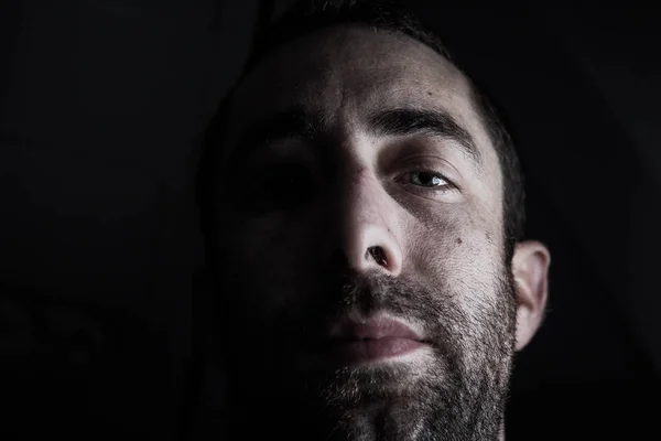 Dramatic portrait of a young man half expressionless face in deep shadow looking at camera isolated on black background.