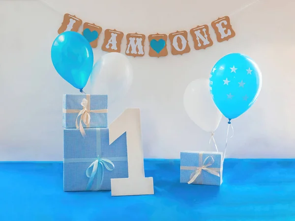 One year birthday decorations set. Decoration for a boy holiday party. Bunch of blue & white balloons, gift boxes, large white kappa board number one shape, and sign with the inscription \