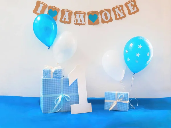 One year birthday decorations set. Decoration for a boy holiday party. Bunch of blue & white balloons, gift boxes, large white kappa board number one shape, and sign with the inscription \