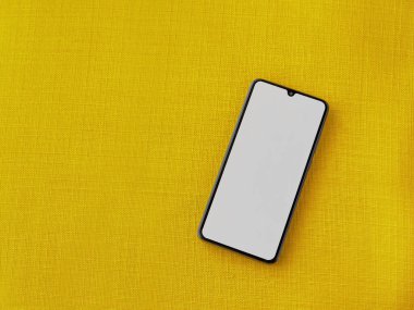 Yüzeyde, sarı bir kumaş zemin üzerinde izole edilmiş boş bir ekran bulunan siyah cep telefonu modeli bulunuyor. Üst görünüm seçici odak ile kapat ve boşluğu kopyala.