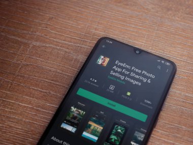 Lod, Israel - 8 Temmuz 2020: EyeEm - Ahşap arka planda siyah bir cep telefonunun ekranında resim paylaşımı ve satışı uygulaması oyun sayfası. Üst görünüm düzlüğü kopyalama alanı ile yatıyordu.