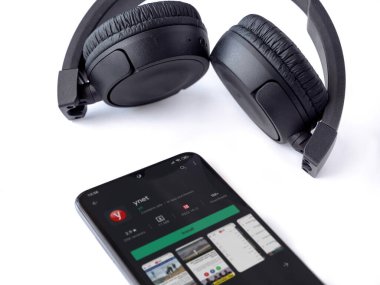 Lod, Israel - 8 Temmuz 2020: Ynet uygulamalı akıllı telefon oyun mağazası sayfası ve beyaz arka planda kablosuz kulaklıklar. Üst görünüm düzlüğü kopyalama alanı ile yatıyordu.