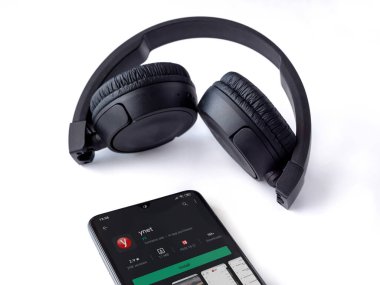 Lod, Israel - 8 Temmuz 2020: Ynet uygulamalı akıllı telefon oyun mağazası sayfası ve beyaz arka planda kablosuz kulaklıklar. Üst görünüm düzlüğü kopyalama alanı ile yatıyordu.