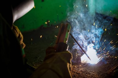 İş yerinde bir kaynakçı. Kıvılcımlar uçuşuyor. İş yerinde maskeli kaynakçı. Kaynakçı mesleği. Etrafta kıvılcımlar var. Kaynak metali. Karanlık arka plan. Parlak ışık.