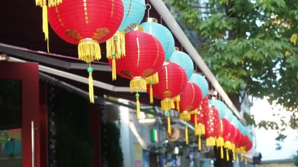 Chinese lanterns sway in the wind in the afternoon. Oriental paper lights of red and blue colors are swaying with the wind on the roof of a building by day on the street — Stock Video