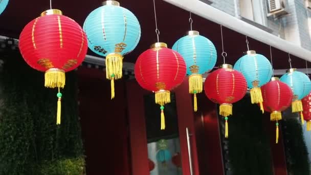 Chinese lantaarns zwaaien in de wind in de middag. Oosterse papier lichten van rode en blauwe kleuren zijn zwaaiend met de wind op het dak van een gebouw door dag op straat — Stockvideo