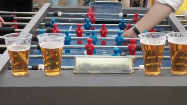 Los jóvenes juegan futbolín y beben cerveza al aire libre. Figuras de futbolistas se mueven a la izquierda y a la derecha jugando futbolín. Trenzas de futbolín. Juego de fútbol de mesa kicker primer plano. Exterior — Vídeo de stock