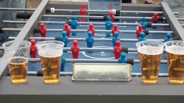 Οι νέοι παίζουν τραπέζι ποδόσφαιρο και ποτό μπύρα σε εξωτερικούς χώρους. Φιγούρες ποδοσφαιριστές μετακίνηση αριστερά και δεξιά άνθρωποι παίζουν επιτραπέζιο ποδοσφαιράκι. Πίνακας Ποδόσφαιρο plaers. Κοντινό τραπέζι Ποδόσφαιρο Ποδόσφαιρο παιχνίδι kicker. Εξωτερική — Αρχείο Βίντεο