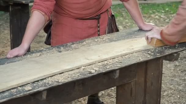 Snickare i medeltida bomullskläder arbeta med en trä med flyg. Man Hews manuellt en träskiva med en plan — Stockvideo