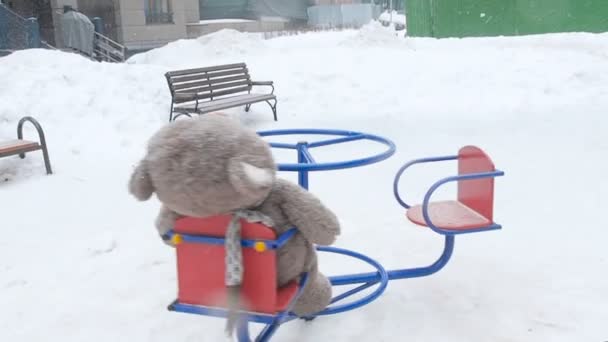 Urso de pelúcia solitário girar sobre o carrossel azul vermelho no playground no inverno durante a queda de neve. Conceito de solidão — Vídeo de Stock