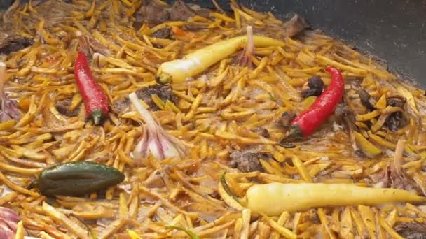 Uzbecká národní pokrm pilaf v velký litinový kotel hoří, nakrájené žluté mrkve, přidán dřišťál, celé chilli papričky česnek a červené zelené a žluté, vroucí, rýže, jehněčí, skopové tuku, — Stock video