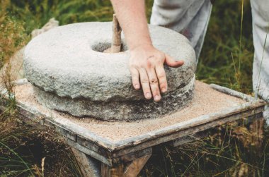 The ancient quern stone hand mill with grain. The man grinds the grain into flour with the help of a millstone. Mens hands on a millstone. Old grinding stones turned by hands clipart