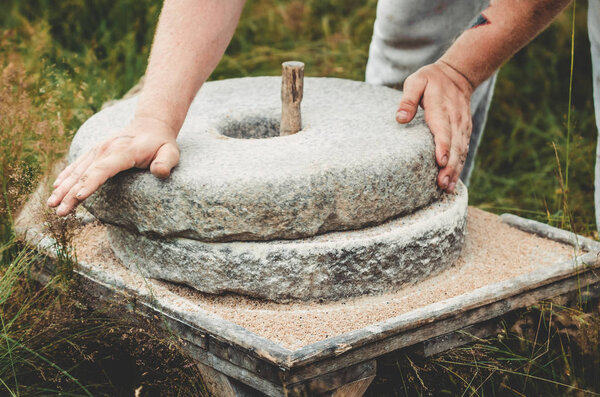 The ancient quern stone hand mill with grain. The man grinds the grain into flour with the help of a millstone. Mens hands on a millstone. Old grinding stones turned by hands