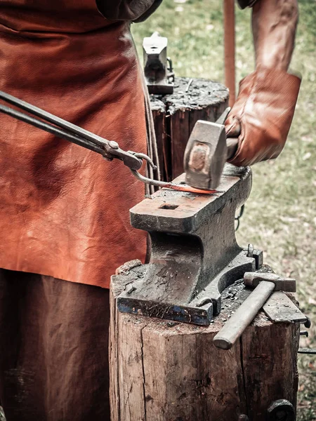The blacksmith in the production process of metal products handmade on the openair workshop. Blacksmith strikes with a hammer on metal. Hands of a blacksmith in leather gloves with red-hot billet