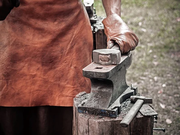 . Mano de herrero con un martillo sobre yunque de acero. Mano de herrero en guantes de cuero con martillo de cerca. El herrero en el proceso de producción de productos metálicos hechos a mano al aire libre — Foto de Stock