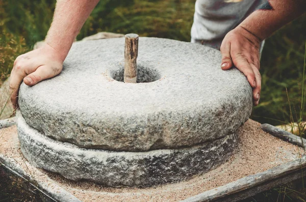 Die alte Quernsteinmühle mit Getreide. Der Mann mahlt das Korn mit Hilfe eines Mühlsteins zu Mehl. Männerhände auf einem Mühlstein. alte Schleifsteine von Hand gedrechselt Stockbild