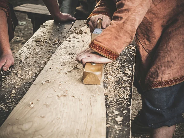 Carpintero en Ropa Medieval de Algodón Trabajando con Madera por Avión. El hombre tala manualmente una tabla de madera con un avión. Construcción de una corteza de madera — Foto de Stock