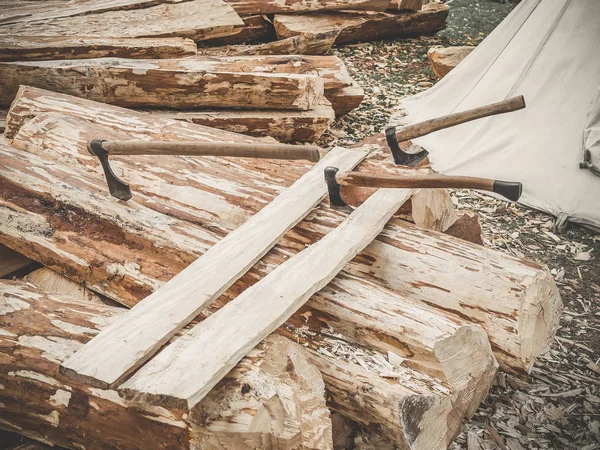 Site in opbouw. Brandhout met assen op een karakter. Veel logs houten plank en drie assen buiten. Bouw van een houten schors. — Stockfoto